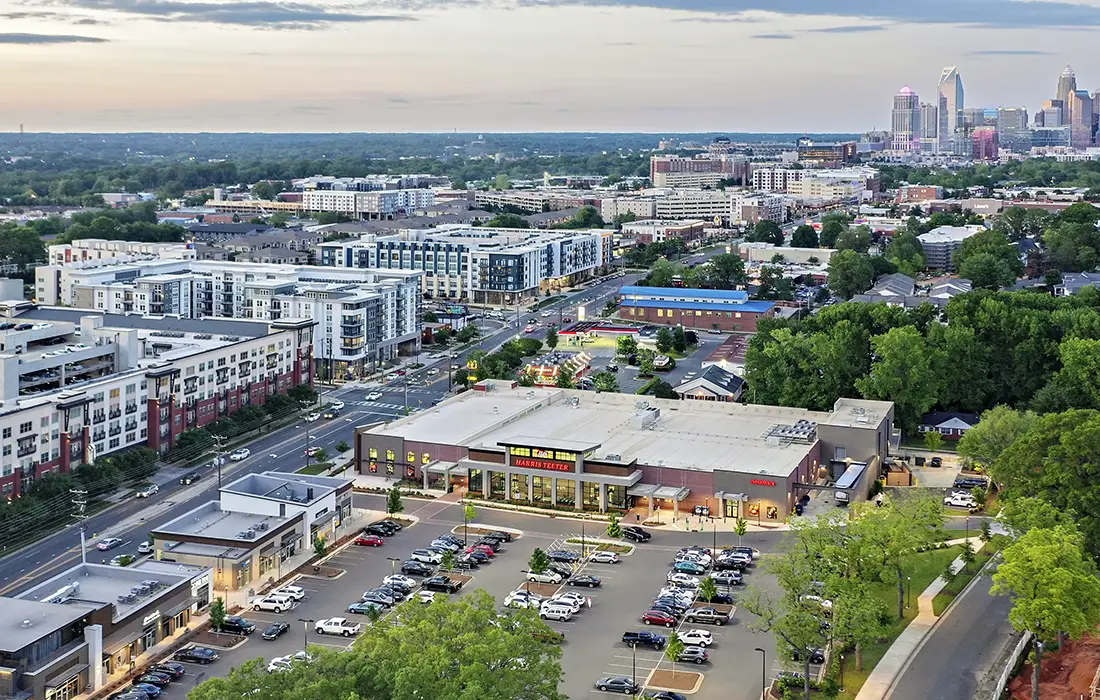 Aerial view of a suburban development with residential buildings and a shopping center, framed by lush greenery, with a city skyline in the distance at dusk.